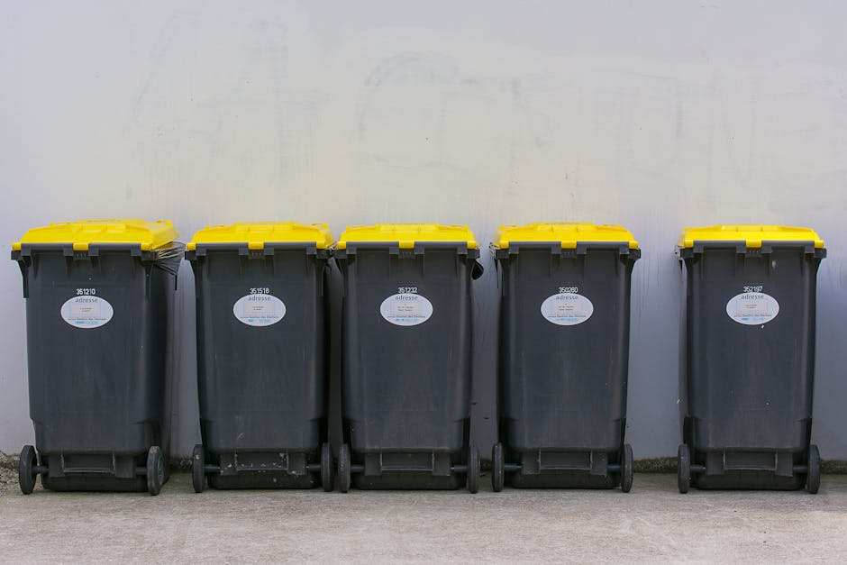 A row of five large wheeled rubbish bins lined up against a plain, light-colored wall. Each bin is made of dark grey plastic with a textured surface, featuring a rectangular shape with slightly rounde